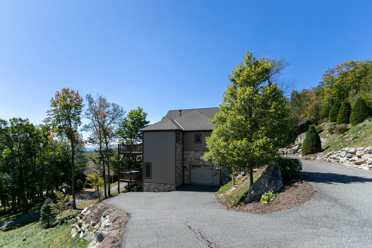A wide exterior shot captures the lodge nestled among mature trees and rocky landscaping. The house sits harmoniously in nature, with inviting porch spaces and the suggestion of tranquil evenings outdoors, possibly after winter adventures.