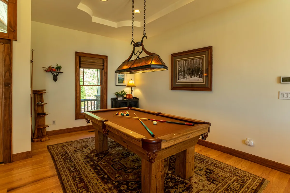 A lively interior scene featuring a classic pool table beneath a chandelier, with a cozy rug underfoot and framed artworks on the wall. Natural light streams through the window, creating a welcoming space for games and socializing.