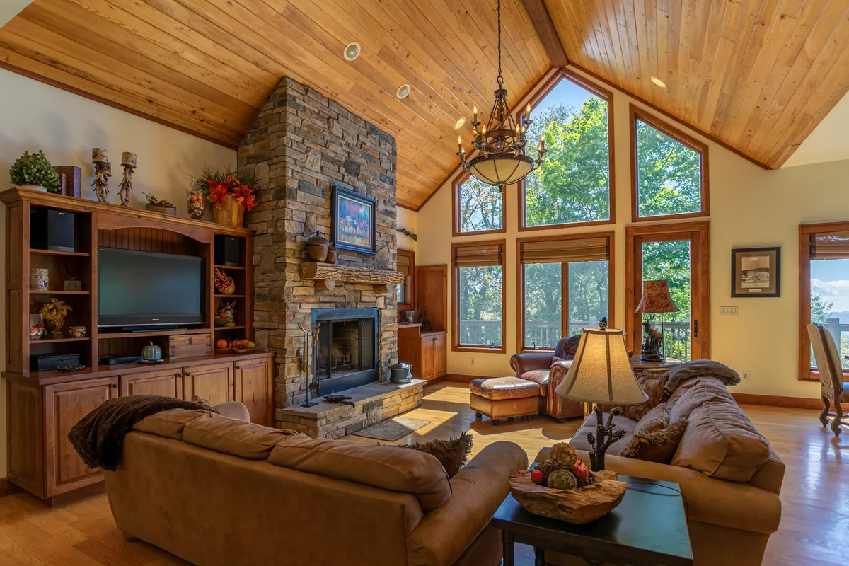 The room is anchored by a prominent stone fireplace, with comfortable seating arranged to encourage conversation and relaxation. Large windows let in ample natural light, while wooden beams and a chandelier add rustic elegance.