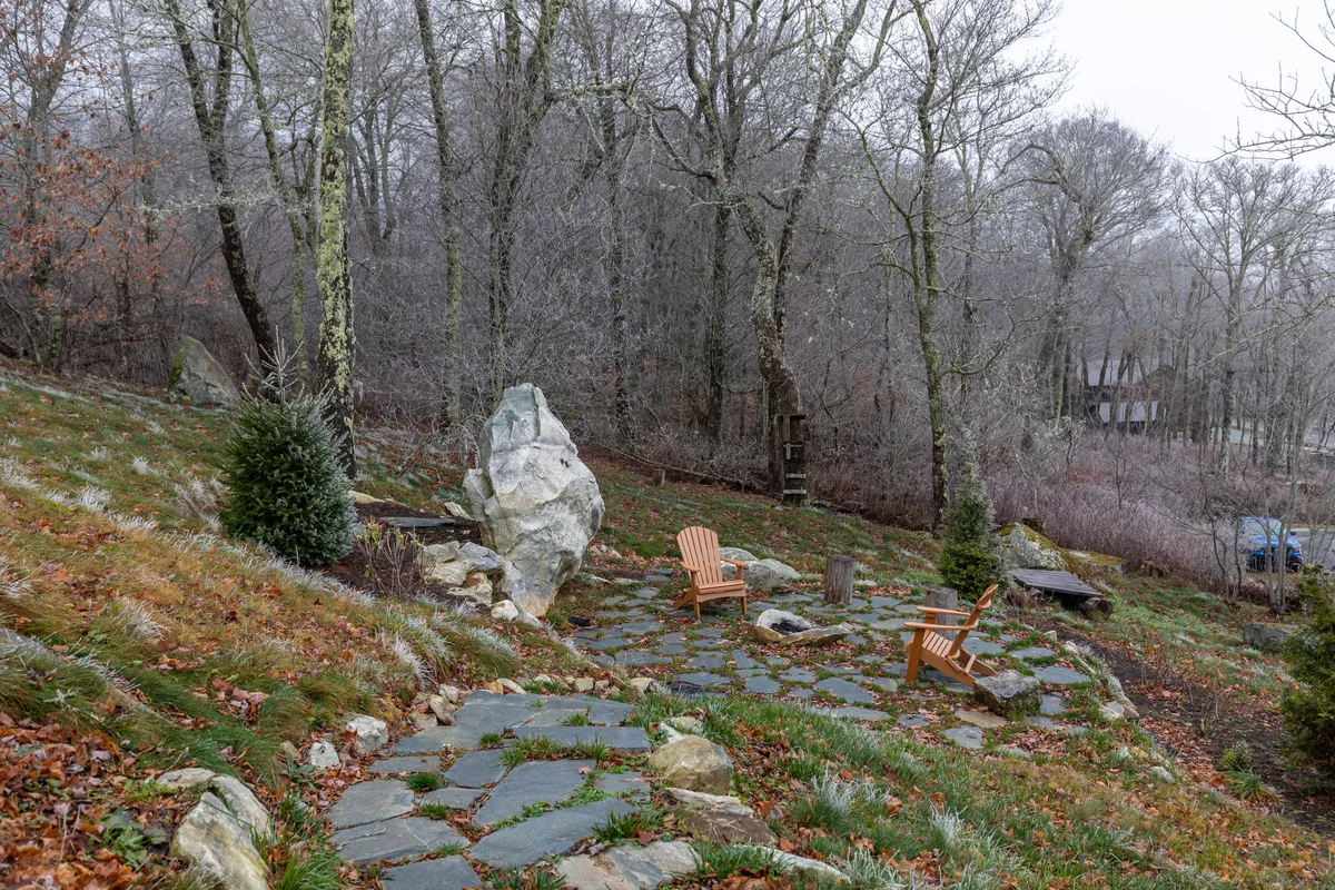 An inviting stone patio features wooden chairs positioned around a large rock, with a tranquil stone pathway set amidst trees and greenery. The scene exudes a sense of relaxation and connection with the outdoors, hinting at activities like stargazing, gathering around a fire, or enjoying a peaceful morning outside.