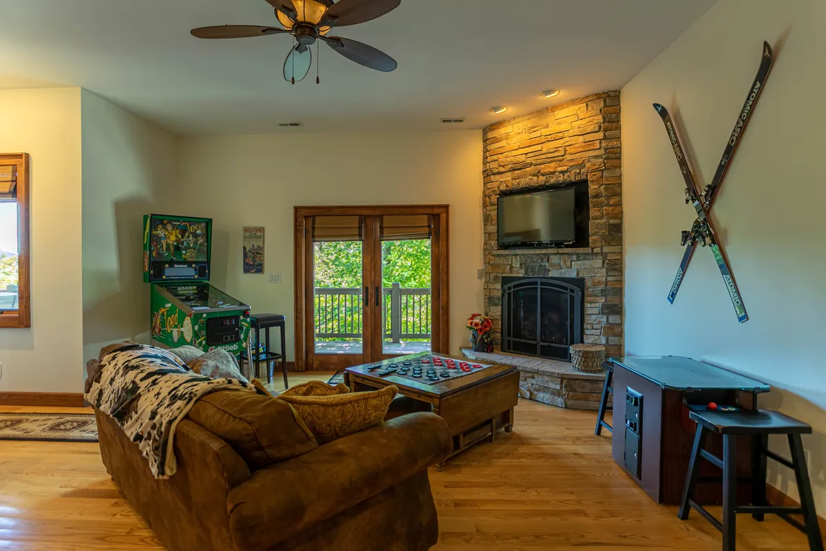 A casual and vibrant game room scene featuring a stone fireplace, TV, comfortable couch, ski-themed decor, and a visible arcade game. The area is set up for both gaming and relaxation, ideal for family game nights or movie marathons.