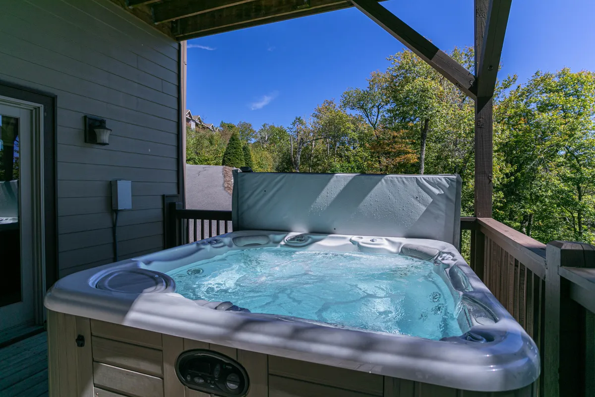 A bubbling hot tub sits on a spacious deck, surrounded by lush greenery. The setting is private and serene, with space for multiple people to relax. The deck area offers a perfect vantage point to take in the mountain environment.