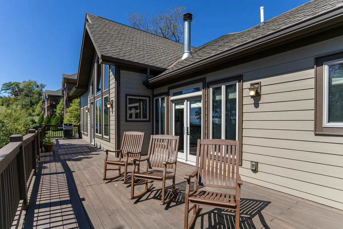 This image captures a tranquil scene on the wooden deck, with comfortable chairs positioned for relaxation and a deck railing that opens to sweeping outdoor views. Large windows hint at a seamless indoor-outdoor connection, while the surrounding natural setting is visible beyond.