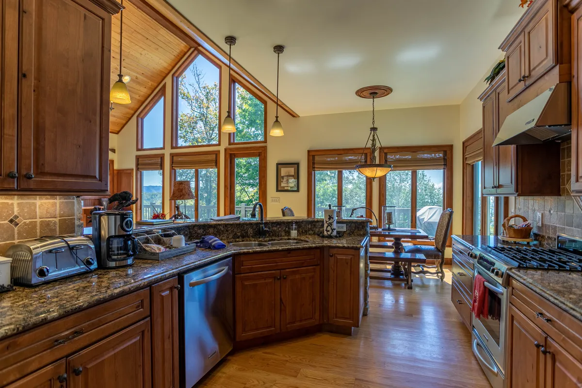 This kitchen and dining area features modern appliances, granite countertops, a dining table bathed in natural light from large windows, and ample space for group meals. The open layout visually connects the kitchen to the rest of the living space.