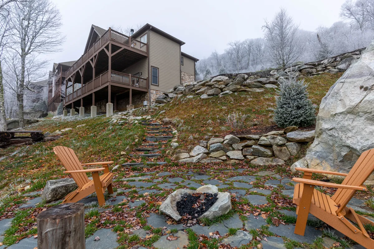 A cozy fire pit surrounded by wooden chairs sits on a stone pathway, with the house on a gentle hill in the background. The area is set up for group gatherings, with a welcoming atmosphere for evening relaxation or fireside treats.