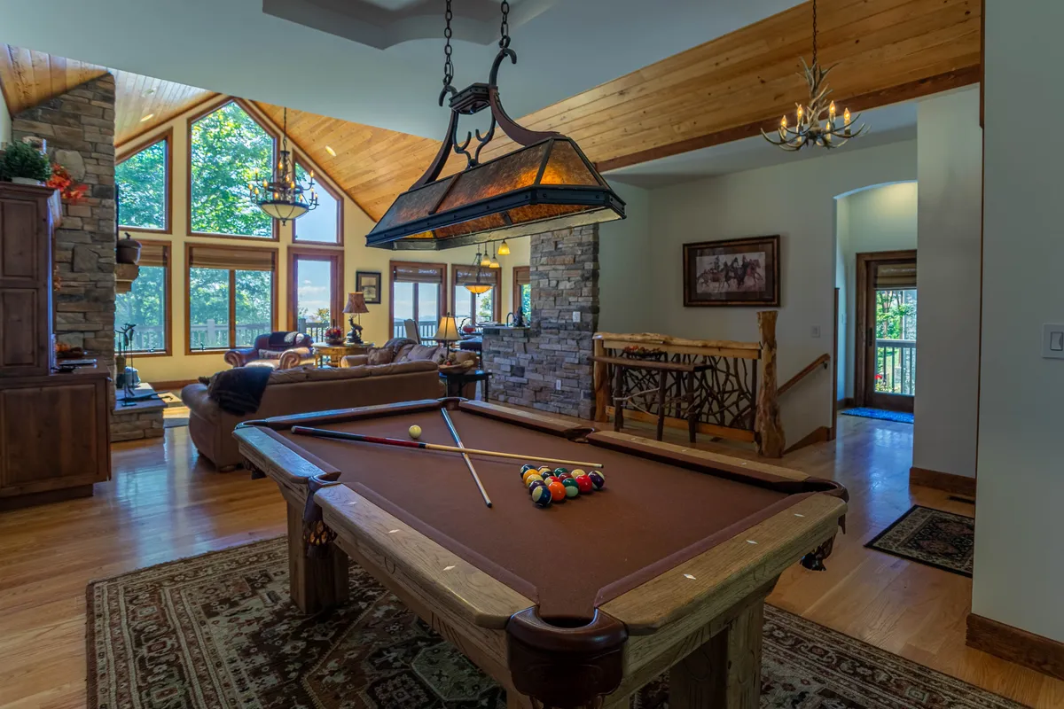 This interior image showcases a welcoming great room featuring a hickory pool table, large windows that frame scenic views, a stone accent wall, wooden beams, and a comfortable seating area. The natural light and expansive windows emphasize a seamless connection to the outdoors, making the room feel bright and airy.