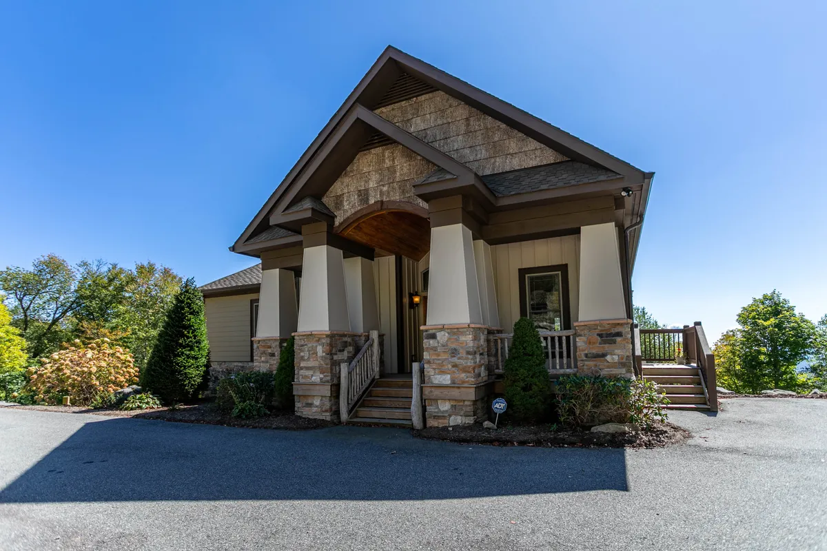 The image showcases the impressive stone facade and charming wooden accents of Heart Rock Lodge's entrance, framed by lush green landscaping. Wide steps lead up to a welcoming porch, perfect for enjoying vibrant sunsets or sipping morning coffee while taking in panoramic mountain views. The inviting exterior hints at the warmth and comfort awaiting inside, making it an ideal setting for family gatherings or outdoor celebrations.